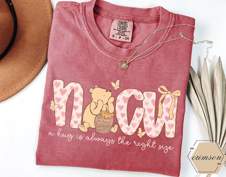 May include: A maroon Comfort Colors t-shirt featuring the word "N CU" in a playful font with heart patterns. A cartoon bear sits beside a honey pot. The text "a hug is always the right size" is below. A gold necklace and a hat are also visible.