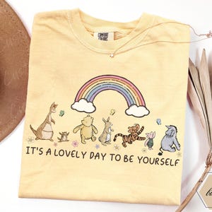 Può includere: Una t-shirt a maniche corte color giallo pallido con una grafica dei personaggi di Winnie the Pooh sotto un arcobaleno. Il testo recita, IT'S A LOVELY DAY TO BE YOURSELF. La maglietta ha un girocollo e un'etichetta Comfort Colors.