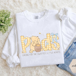 May include: White sweatshirt with the word "Pads" in yellow plaid letters, featuring a cartoon bear and honey pot. The text "Caring for the sweetest little hearts" is below the word "Pads". A pair of blue jeans is visible.