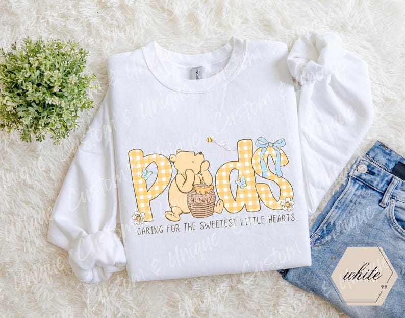 May include: White sweatshirt with the word "Pads" in yellow plaid letters, featuring a cartoon bear and honey pot. The text "Caring for the sweetest little hearts" is below the word "Pads". A pair of blue jeans is visible.