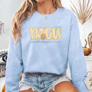 May include: Light blue sweatshirt with the text "NICU" in yellow gingham letters, featuring a cartoon bear and honey pot design. The words "neonatal intensive care unit" are below the main text.
