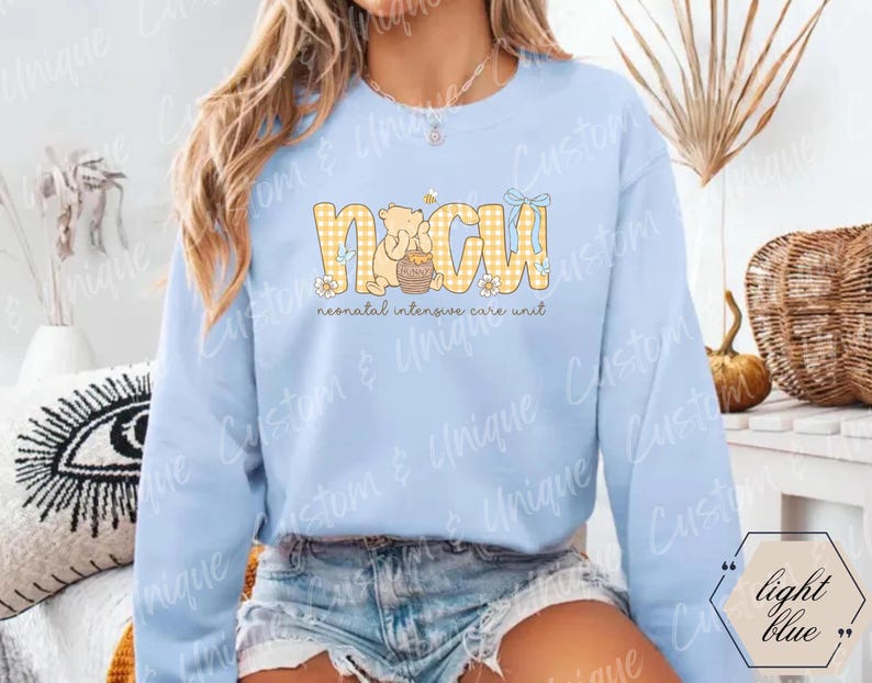 May include: Light blue sweatshirt with the text "NICU" in yellow gingham letters, featuring a cartoon bear and honey pot design. The words "neonatal intensive care unit" are below the main text.