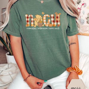 May include: Sage green t-shirt with the letters "NICU" in a plaid design, featuring a cartoon bear and pumpkin graphic. The text "neonatal intensive care unit" is below the letters. The shirt is a casual, comfortable style.