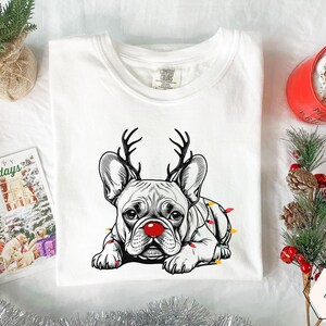 May include: White t-shirt featuring a black and white illustration of a French Bulldog with reindeer antlers and a red nose. The dog is adorned with colorful Christmas lights. A holiday card and a red mug are in the background.