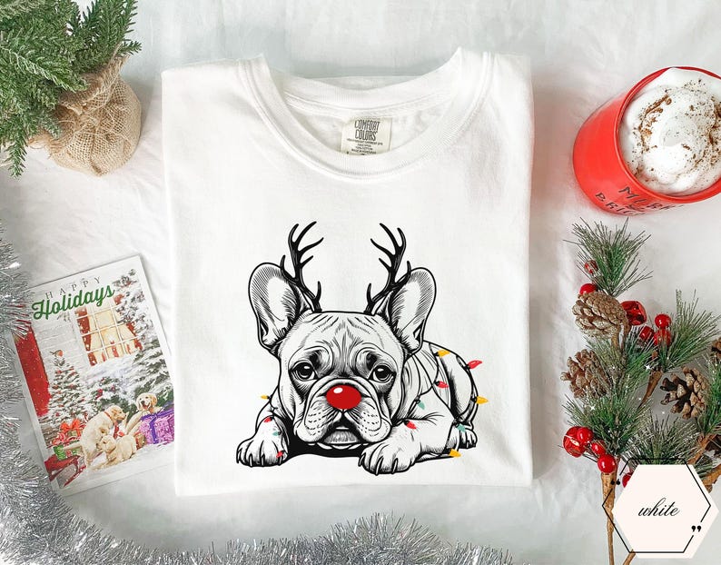 May include: White t-shirt featuring a black and white illustration of a French Bulldog with reindeer antlers and a red nose. The dog is adorned with colorful Christmas lights. A holiday card and a red mug are in the background.