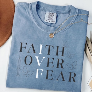 Puede incluir: Una camiseta Comfort Colors azul claro con las palabras "FAITH OVER FEAR" impresas en blanco y negro. La camiseta presenta un gráfico de jeringa y mariposa. Un collar de oro con un colgante descansa sobre la camiseta.