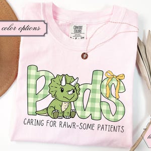 May include: A pale pink t-shirt featuring the word "Peds" in green gingham letters and a cartoon dinosaur. The text "Caring for Rawr-some Patients" is below. A gold necklace and a brown hat are also visible.