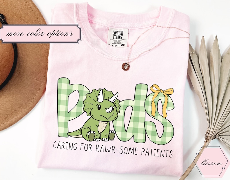 May include: A pale pink t-shirt featuring the word "Peds" in green gingham letters and a cartoon dinosaur. The text "Caring for Rawr-some Patients" is below. A gold necklace and a brown hat are also visible.