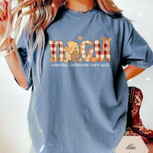 May include: Blue jean colored t-shirt with the letters "NICU" in a plaid pattern, with a Winnie the Pooh graphic and the words "neonatal intensive care unit". The shirt has short sleeves.
