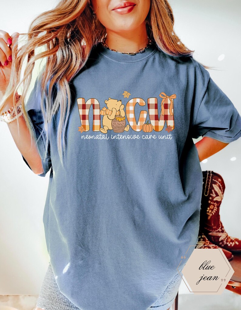 May include: Blue jean colored t-shirt with the letters "NICU" in a plaid pattern, with a Winnie the Pooh graphic and the words "neonatal intensive care unit". The shirt has short sleeves.