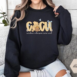 May include: Black crew neck sweatshirt featuring the text "CICU cardiac intensive care unit" in a yellow gingham design. The design includes a cartoon bear holding a honey pot, flowers, and butterflies. A comfortable, casual style.