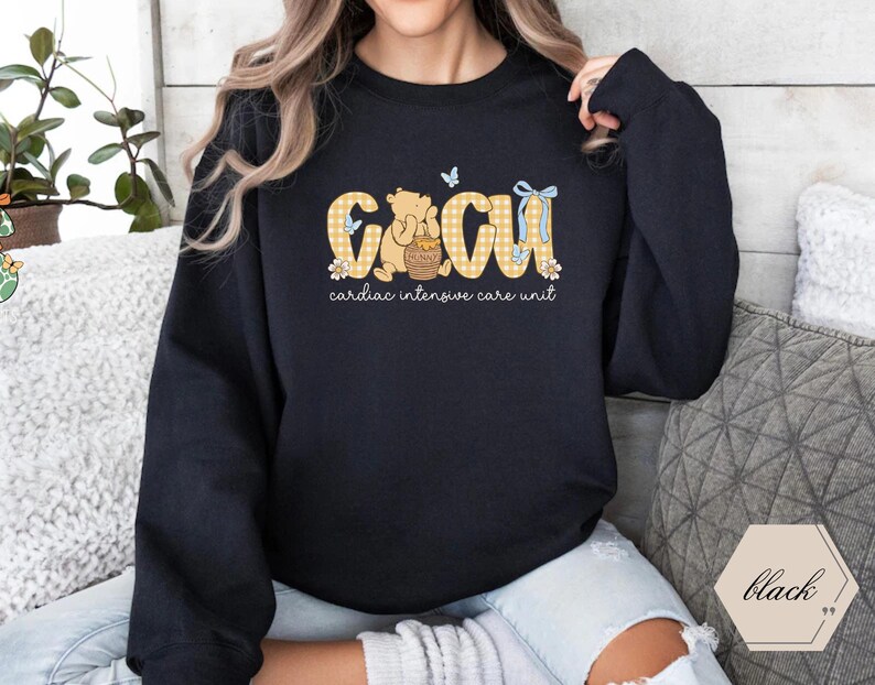 May include: Black crew neck sweatshirt featuring the text "CICU cardiac intensive care unit" in a yellow gingham design. The design includes a cartoon bear holding a honey pot, flowers, and butterflies. A comfortable, casual style.