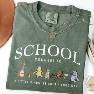 May include: A moss green short-sleeve t-shirt featuring the white text SCHOOL COUNSELOR and a graphic of Winnie the Pooh characters. Below the characters, the text reads A LITTLE KINDNESS GOES A LONG WAY. The shirt has a Comfort Colors brand label.