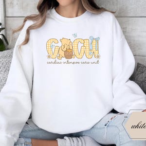 May include: White sweatshirt with a graphic design. The design features the letters "CIU" in yellow plaid, with a cartoon bear holding a honey pot. The text "cardiac intensive care unit" is below the letters.
