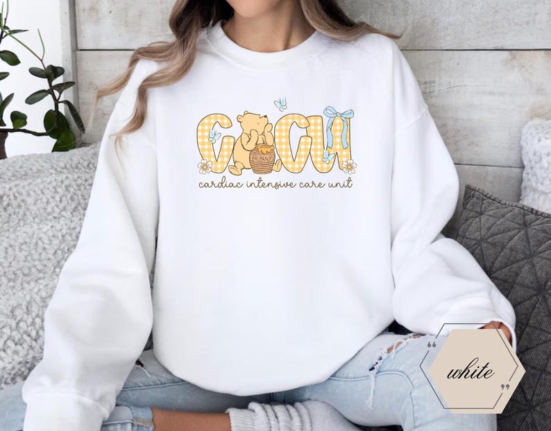 May include: White sweatshirt with a graphic design. The design features the letters "CIU" in yellow plaid, with a cartoon bear holding a honey pot. The text "cardiac intensive care unit" is below the letters.