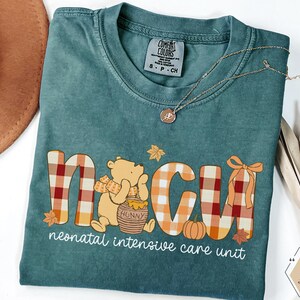 May include: Teal Comfort Colors t-shirt with a Winnie the Pooh-themed design. The design includes the letters "NICU" in a plaid pattern, a pumpkin, and the words "neonatal intensive care unit". A gold necklace and a brown hat are also visible.