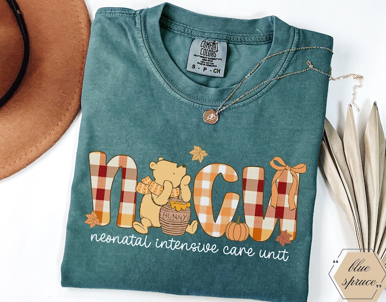 May include: Teal Comfort Colors t-shirt with a Winnie the Pooh-themed design. The design includes the letters "NICU" in a plaid pattern, a pumpkin, and the words "neonatal intensive care unit". A gold necklace and a brown hat are also visible.
