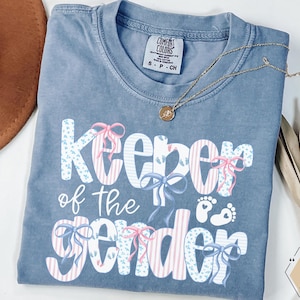 May include: A blue t-shirt with the words "Keeper of the Gender" in white, decorated with bows and baby footprints. A gold necklace rests on the shirt. The shirt is size S.