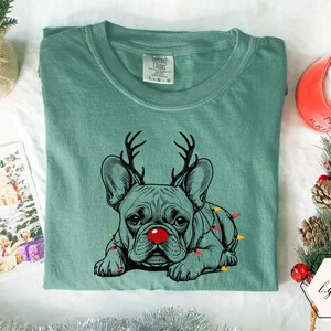 May include: A seafoam green t-shirt featuring a graphic of a French Bulldog with reindeer antlers and a red nose. The dog is adorned with Christmas lights. A holiday card and a mug of hot chocolate are in the background.