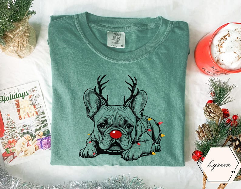 May include: A seafoam green t-shirt featuring a graphic of a French Bulldog with reindeer antlers and a red nose. The dog is adorned with Christmas lights. A holiday card and a mug of hot chocolate are in the background.