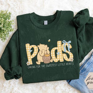 May include: A forest green sweatshirt with the word "Pods" in yellow gingham letters, featuring a cartoon bear and honey pot. The text "Caring for the sweetest little hearts" is below the graphic. A small bee and blue bow are also included.
