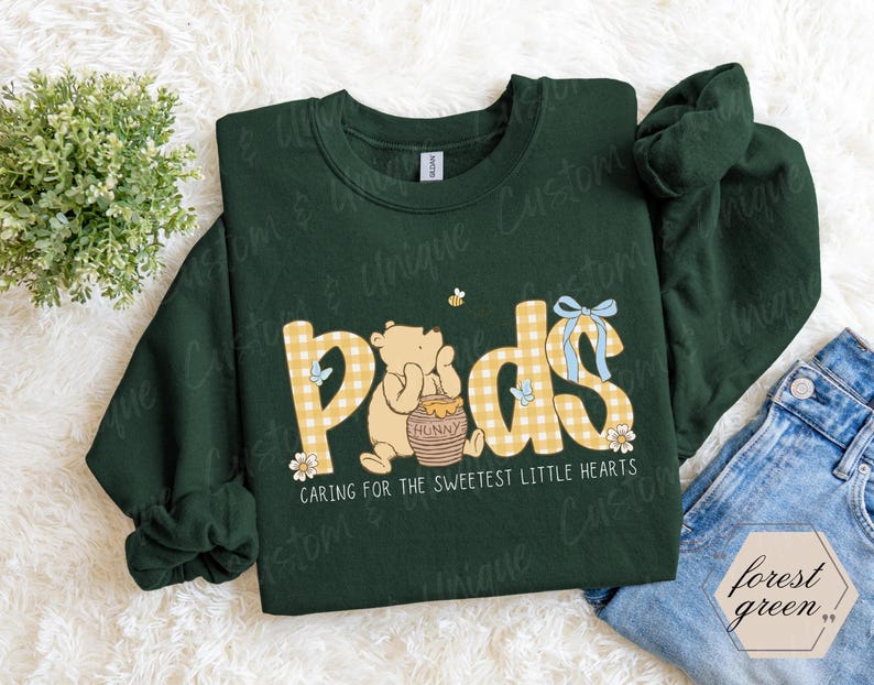 May include: A forest green sweatshirt with the word "Pods" in yellow gingham letters, featuring a cartoon bear and honey pot. The text "Caring for the sweetest little hearts" is below the graphic. A small bee and blue bow are also included.