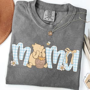 May include: Gray Comfort Colors t-shirt with the word "mama" in a blue gingham pattern, featuring a Winnie the Pooh graphic. A gold necklace and a brown hat are also visible.