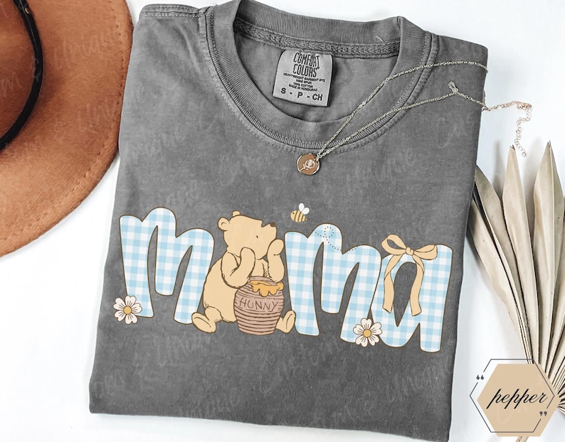 May include: Gray Comfort Colors t-shirt with the word "mama" in a blue gingham pattern, featuring a Winnie the Pooh graphic. A gold necklace and a brown hat are also visible.