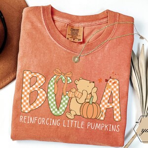 Könnte beinhalten: Orangefarbenes Comfort Colors T-Shirt mit einer Grafik eines Bären, eines Kürbisses und dem Text "BCA Reinforcing Little Pumpkins". Die Buchstaben B, C und A sind in orange- und grün karierten Mustern gehalten. Eine goldene Halskette liegt über dem Shirt.