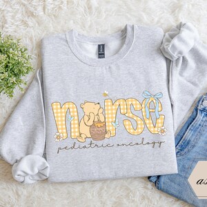 Peut inclure: Un sweat-shirt gris clair avec le mot "nurse" en lettres &agrave; carreaux jaunes, repr&eacute;sentant un ours de dessin anim&eacute; et un pot de miel. Le texte "pediatric oncology" est en dessous du mot "nurse". Un n&oelig;ud bleu et une abeille sont &eacute;galement pr&eacute;sents.