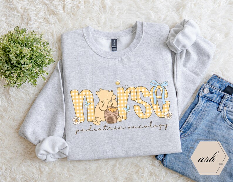 Peut inclure: Un sweat-shirt gris clair avec le mot "nurse" en lettres &agrave; carreaux jaunes, repr&eacute;sentant un ours de dessin anim&eacute; et un pot de miel. Le texte "pediatric oncology" est en dessous du mot "nurse". Un n&oelig;ud bleu et une abeille sont &eacute;galement pr&eacute;sents.