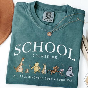 May include: A teal short-sleeved t-shirt with the text SCHOOL COUNSELOR and A LITTLE KINDNESS GOES A LONG WAY. The graphic features illustrations of various cartoon animals including a bear, tiger, and donkey. A gold-toned necklace rests on the shirt.