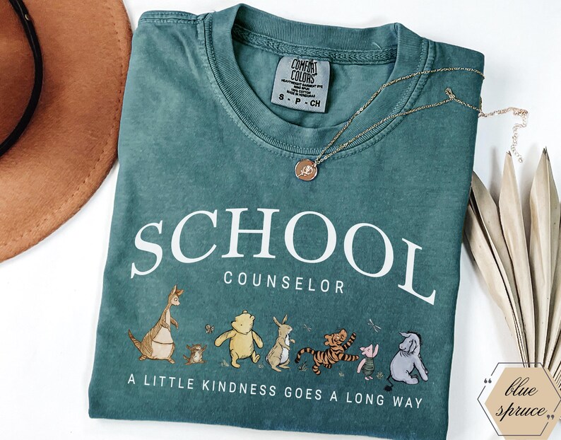 May include: A teal short-sleeved t-shirt with the text SCHOOL COUNSELOR and A LITTLE KINDNESS GOES A LONG WAY. The graphic features illustrations of various cartoon animals including a bear, tiger, and donkey. A gold-toned necklace rests on the shirt.