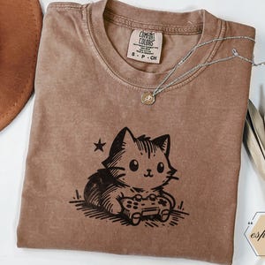 May include: Espresso-colored t-shirt featuring a black graphic of a cat playing a video game. The shirt has a crew neck and short sleeves. A silver necklace with a small pendant rests on the shirt. The shirt has a Comfort Colors tag.