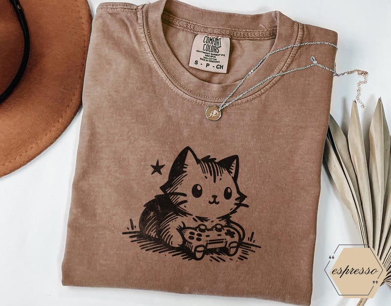 May include: Espresso-colored t-shirt featuring a black graphic of a cat playing a video game. The shirt has a crew neck and short sleeves. A silver necklace with a small pendant rests on the shirt. The shirt has a Comfort Colors tag.