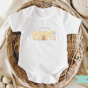 May include: White baby bodysuit featuring the text "I love my Mimi". The design includes a bear and honey pot. The text is in a playful font. The bodysuit has short sleeves and popper fastenings at the bottom.