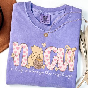 May include: A lilac Comfort Colors t-shirt featuring the word "N CUH" in pink letters with heart patterns. A cartoon bear sits beside a honey pot. The text "a hug is always the right size" is below the graphic.
