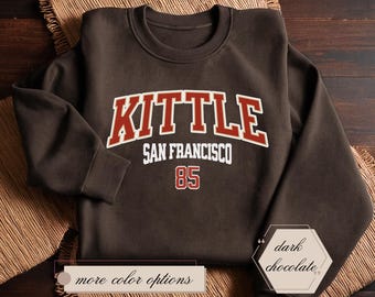 Può includere: Felpa marrone cioccolato scuro con il testo "KITTLE SAN FRANCISCO 85" in lettere rosse e bianche.
