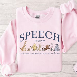 May include: Light pink sweatshirt with the words "SPEECH THERAPY" in navy blue arched across the top. Below the text are cartoon characters. The text "EVERY WAY TO COMMUNICATE IS THE RIGHT WAY" is printed below the characters.