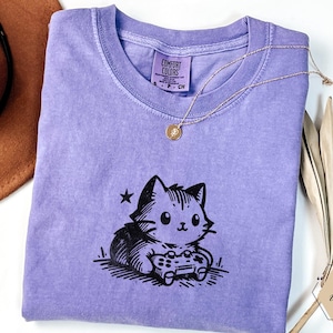 May include: A violet Comfort Colors t-shirt featuring a black and white cartoon cat playing a video game. A gold necklace with a pendant rests on the shirt. A brown hat and dried palm leaves are in the background.