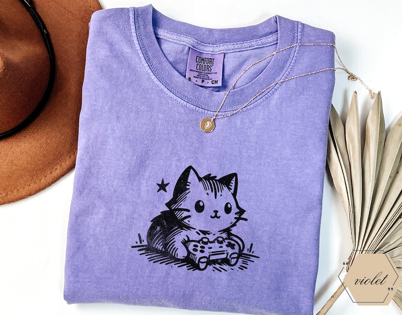 May include: A violet Comfort Colors t-shirt featuring a black and white cartoon cat playing a video game. A gold necklace with a pendant rests on the shirt. A brown hat and dried palm leaves are in the background.