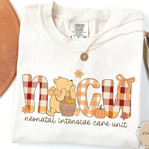 May include: Cream-colored t-shirt with the letters NICU in a plaid pattern, featuring a cartoon bear, a honey pot, and a pumpkin. The text "neonatal intensive care unit" is printed below the letters. A gold necklace is draped on the shirt.
