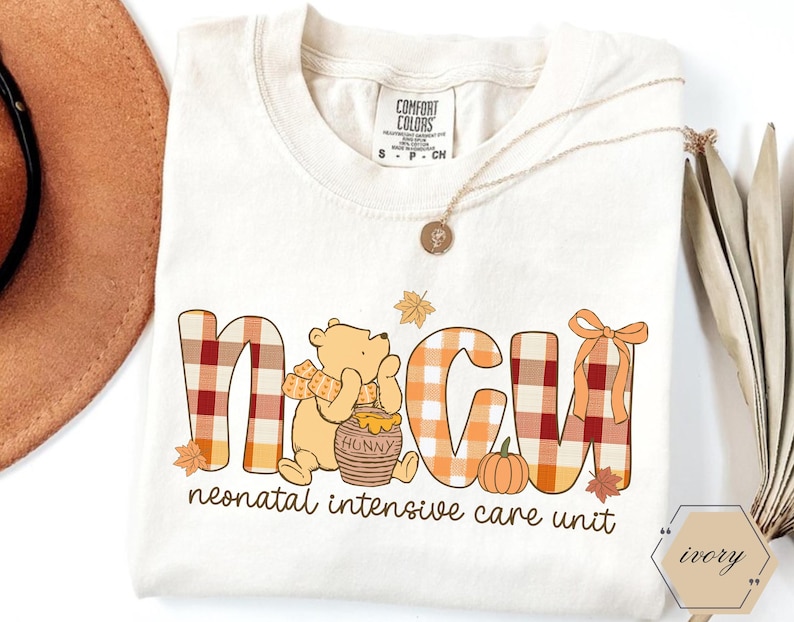 May include: Cream-colored t-shirt with the letters NICU in a plaid pattern, featuring a cartoon bear, a honey pot, and a pumpkin. The text "neonatal intensive care unit" is printed below the letters. A gold necklace is draped on the shirt.