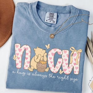 May include: A blue Comfort Colors t-shirt featuring a graphic of Winnie the Pooh with the word "NCU" in pink letters with heart patterns. The shirt also includes the phrase "a hug is always the right size". A gold necklace and decorative elements are also visible.