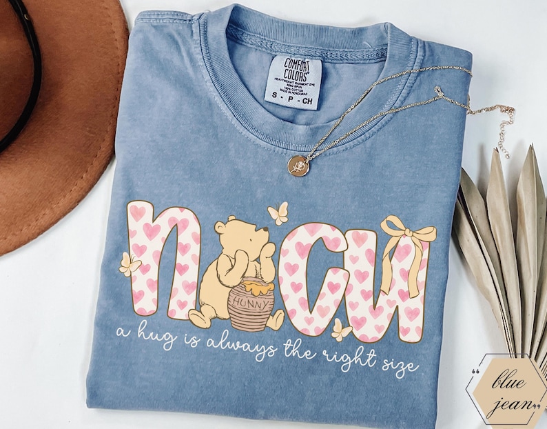 May include: A blue Comfort Colors t-shirt featuring a graphic of Winnie the Pooh with the word "NCU" in pink letters with heart patterns. The shirt also includes the phrase "a hug is always the right size". A gold necklace and decorative elements are also visible.