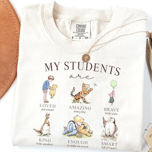 May include: Cream-colored t-shirt with the text "MY STUDENTS are" and illustrations of Winnie the Pooh characters. The shirt features the words "LOVED," "AMAZING," "BRAVE," "KIND," "ENOUGH," and "SMART."