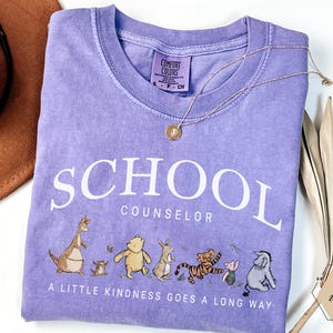 May include: A violet short-sleeved t-shirt featuring the white text SCHOOL COUNSELOR and the phrase A LITTLE KINDNESS GOES A LONG WAY. Below the text are illustrations of Winnie the Pooh characters including Roo, Pooh, Rabbit, Tigger, Piglet, and Eeyore.