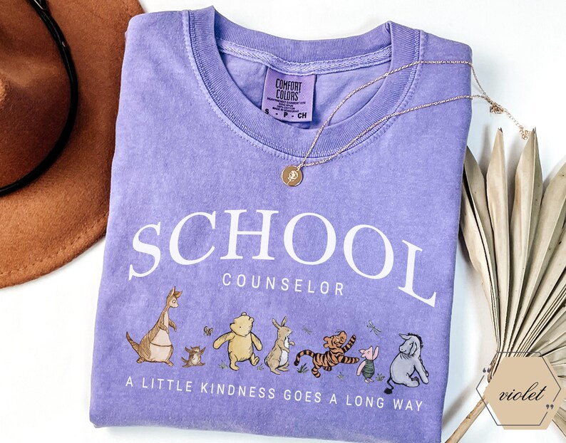 May include: A violet short-sleeved t-shirt featuring the white text SCHOOL COUNSELOR and the phrase A LITTLE KINDNESS GOES A LONG WAY. Below the text are illustrations of Winnie the Pooh characters including Roo, Pooh, Rabbit, Tigger, Piglet, and Eeyore.