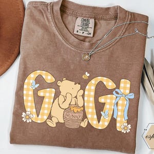 May include: Brown t-shirt with the word "GIGI" in yellow gingham print, featuring a cartoon bear, honey pot, flowers, and butterflies. A silver necklace with a pendant rests on the shirt. The shirt has a Comfort Colors tag.