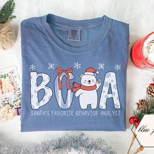 Op de afbeelding: Blauw spijkerbroekkleurig T-shirt met de witte tekst "BOCA", met een cartoon ijsbeer met een kerstmuts en sjaal. De tekst "SANTA'S FAVORITE BEHAVIOR ANALYST" staat eronder. Het shirt heeft een ronde hals en korte mouwen.
